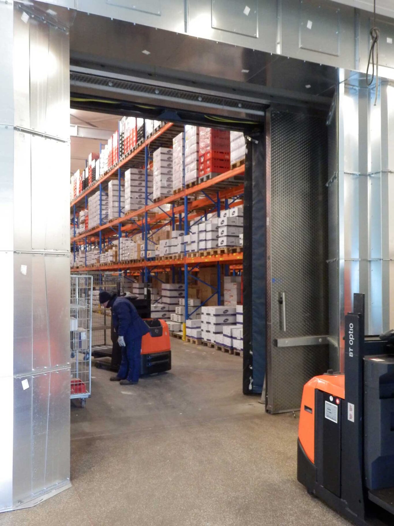 Masterveil PowerStream DSB Comfort luftridå installerad vid en stor frysdörr. Inuti frysen står en arbetare framför sin lastbil och bär varma kläder för att bekämpa den kalla temperaturen. Luftridån hjälper till att behålla kylan inne i frysen samtidigt som den möjliggör enkel åtkomst för personal och utrustning.
