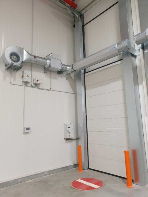 Masterveil AS-K air curtain above a sectional freezer door with circular duct and fan to keep cold air inside and warm air outside.